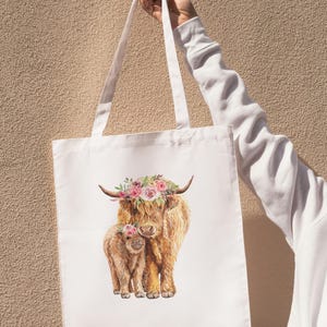May include: White tote bag featuring a watercolor illustration of a Highland cow and calf, both adorned with floral crowns. The bag has long white handles and is held up against a textured beige background.