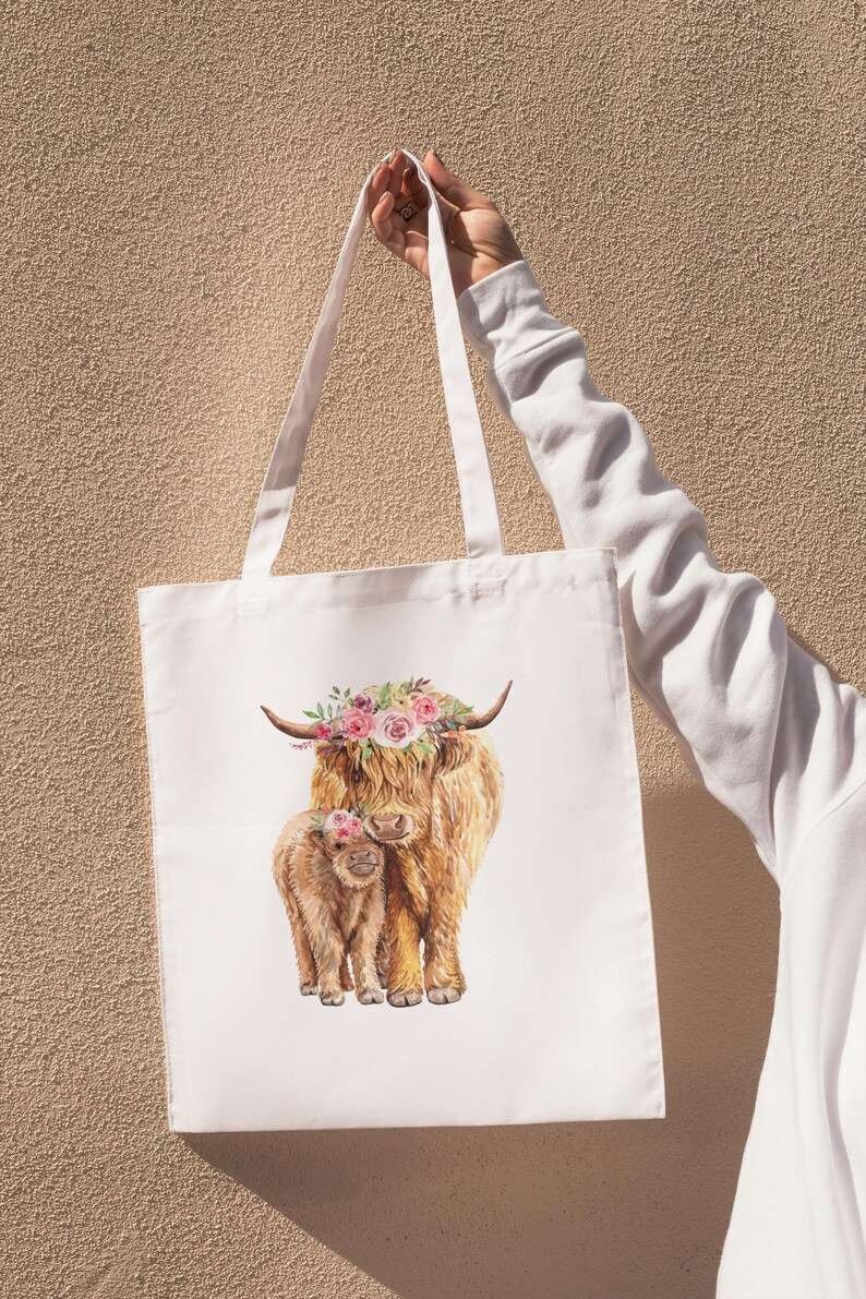 May include: White tote bag featuring a watercolor illustration of a Highland cow and calf, both adorned with floral crowns. The bag has long white handles and is held up against a textured beige background.