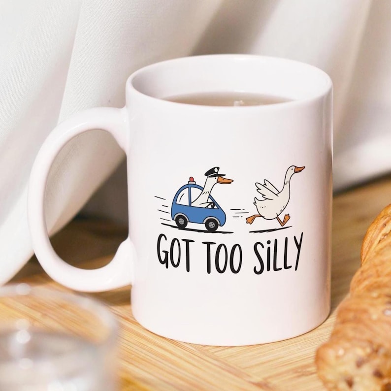 May include: White ceramic mug with a whimsical design. The mug features a cartoon goose in a police car chasing another goose. The text "GOT TOO SILLY" is printed below the illustration. A fun and playful gift.