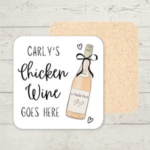 Op de afbeelding: Een vierkante witte onderzetter met de tekst "CARLY'S Chicken Wine GOES HERE". De onderzetter heeft een illustratie van een wijnfles met een zwarte strik en de woorden "La Vieille Ferme". Achter de onderzetter bevindt zich een tweede, rechthoekige kurken onderzetter.