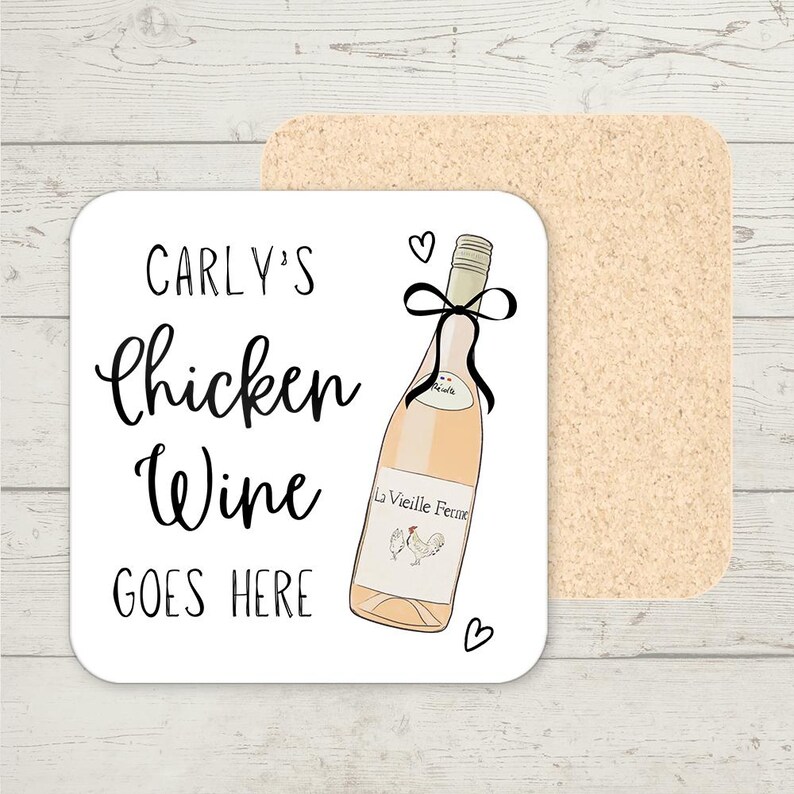 Op de afbeelding: Een vierkante witte onderzetter met de tekst "CARLY'S Chicken Wine GOES HERE". De onderzetter heeft een illustratie van een wijnfles met een zwarte strik en de woorden "La Vieille Ferme". Achter de onderzetter bevindt zich een tweede, rechthoekige kurken onderzetter.