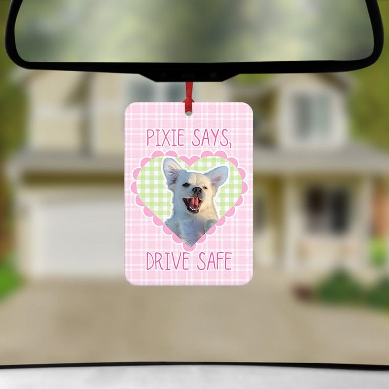 May include: A rectangular car air freshener featuring a pink and white checkered background. A central green and white checkered heart frame contains a photograph of a white dog with its mouth open. The text reads PIXIE SAYS, DRIVE SAFE.