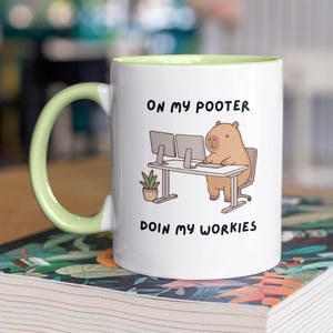 May include: White ceramic mug with a light green handle and interior. The mug features a cartoon capybara at a desk with two computer monitors. Text on the mug reads "ON MY POOTER DOIN MY WORKIES."