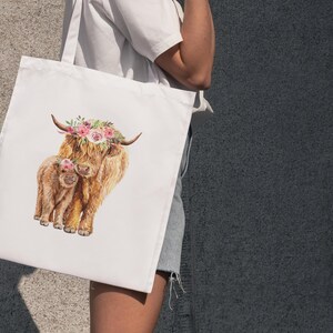 May include: White tote bag featuring a watercolor illustration of a Highland cow and calf. The adult cow wears a floral crown. The artwork is in shades of brown, tan, and pink, with a focus on the animals' fluffy fur.