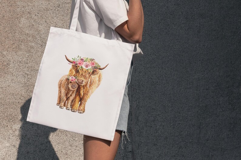 May include: White tote bag featuring a watercolor illustration of a Highland cow and calf. The adult cow wears a floral crown. The artwork is in shades of brown, tan, and pink, with a focus on the animals' fluffy fur.