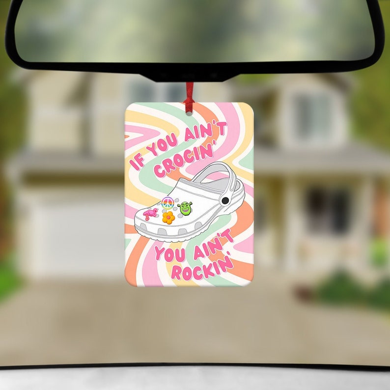 May include: A pink and white car air freshener with a white clog shoe with colourful charms. The text on the air freshener reads "If you ain't crocin' you ain't rockin'"