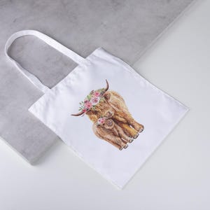 May include: White tote bag featuring a watercolor illustration of a Highland cow and calf adorned with floral crowns. The bag has a simple design with a rectangular shape and two white handles. The artwork is in shades of brown, pink, and green.