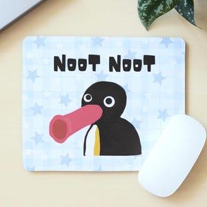 May include: A rectangular mouse mat with a cartoon penguin design, featuring a pink beak and the words "NOOT NOOT". The background is light blue with white star patterns.
