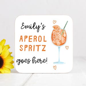 May include: A square white coaster featuring the black text Emily's and goes here! in a script font, with the orange text APEROL SPRITZ in a block font. An illustration of an orange cocktail in a stemmed glass with a green straw and hearts.