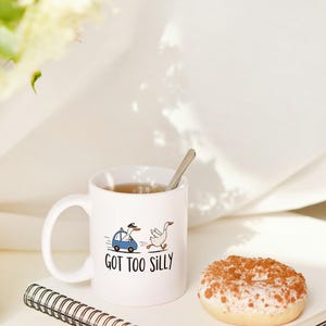 May include: White ceramic mug with the phrase "GOT TOO SILLY" and a cartoon of a goose in a police car chasing another goose. A donut with white frosting and brown sprinkles sits next to the mug. A notebook and spoon are also visible.