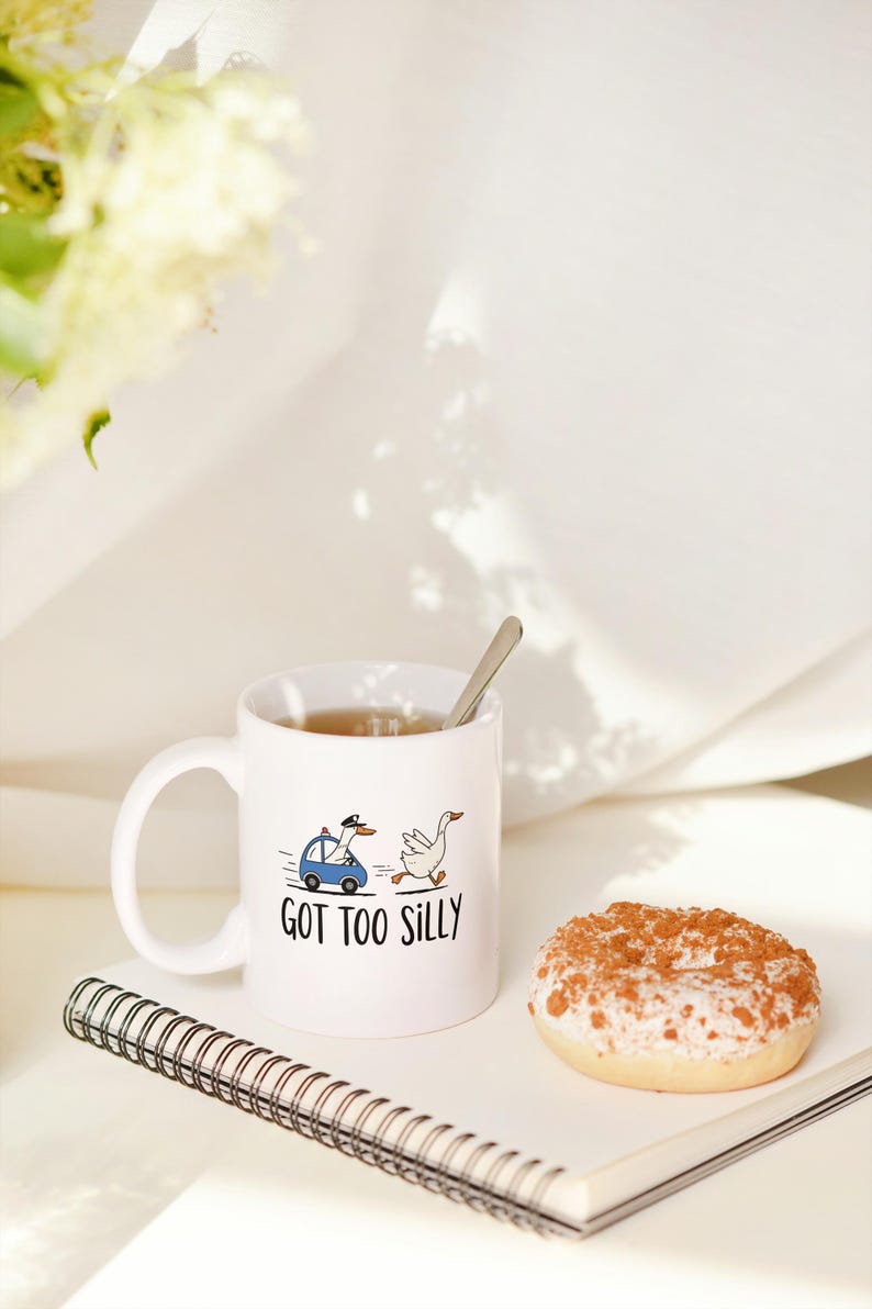 May include: White ceramic mug with the phrase "GOT TOO SILLY" and a cartoon of a goose in a police car chasing another goose. A donut with white frosting and brown sprinkles sits next to the mug. A notebook and spoon are also visible.