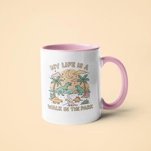 May include: White ceramic mug with a pink interior and handle. The mug features a colorful illustration of dinosaurs in a park setting with the text "MY LIFE IS A WALK IN THE PARK".