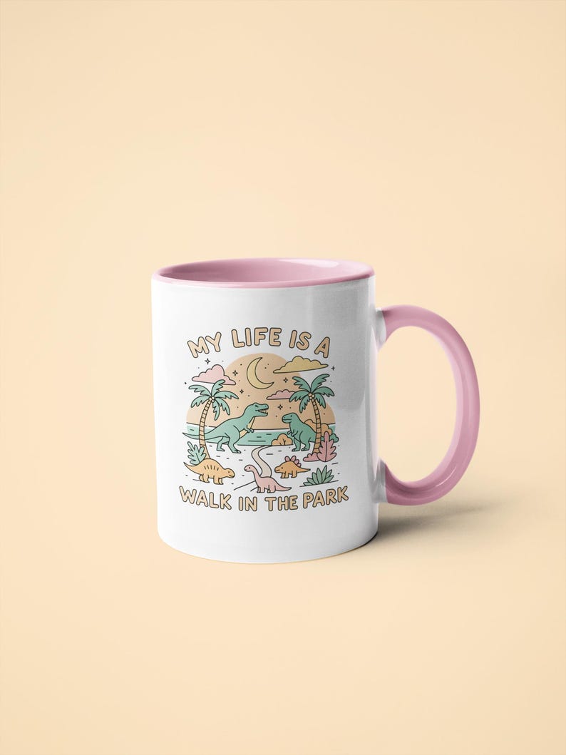 May include: White ceramic mug with a pink interior and handle. The mug features a colorful illustration of dinosaurs in a park setting with the text "MY LIFE IS A WALK IN THE PARK".