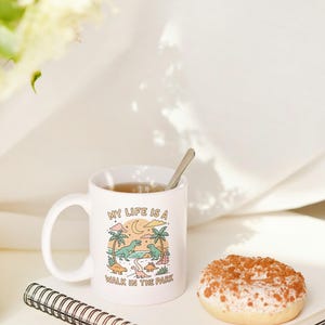 May include: White ceramic mug with the text "MY LIFE IS A WALK IN THE PARK" and a cartoon dinosaur scene. A spoon rests in the mug filled with tea. A glazed donut and a spiral notebook are also in the image.