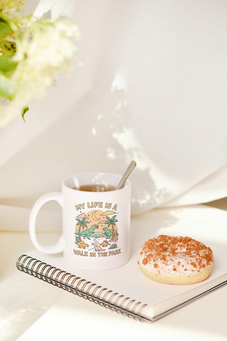 May include: White ceramic mug with the text "MY LIFE IS A WALK IN THE PARK" and a cartoon dinosaur scene. A spoon rests in the mug filled with tea. A glazed donut and a spiral notebook are also in the image.