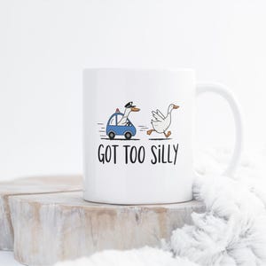 May include: White ceramic mug featuring a cartoon goose in a police car chasing another goose. The mug has the text "GOT TOO SILLY" in black. The mug is on a wooden surface with a white fluffy blanket in the background.