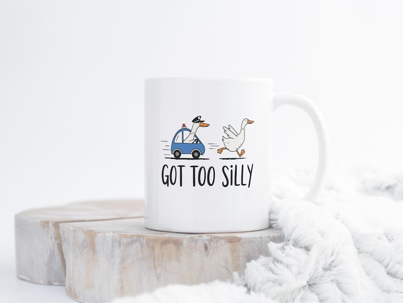 May include: White ceramic mug featuring a cartoon goose in a police car chasing another goose. The mug has the text "GOT TOO SILLY" in black. The mug is on a wooden surface with a white fluffy blanket in the background.