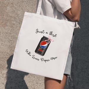 May include: White tote bag featuring a Pepsi Max can design with a pink bow. The text reads "Just a Girl Who Loves Pepsi Max" in black script. The bag has two handles.