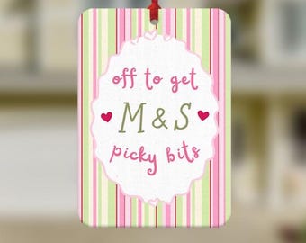 May include: A rectangular hanging air freshener featuring a vertical striped pattern in shades of pink, light green, and white. A central white scalloped label displays the pink and olive green text "off to get M&S picky bits" with two small red hearts.