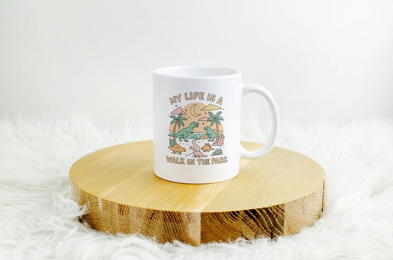 May include: White ceramic mug with a whimsical dinosaur design. The mug features a colorful illustration of dinosaurs, palm trees, and the text "MY LIFE IS A WALK IN THE PARK". The mug is placed on a wooden round surface.