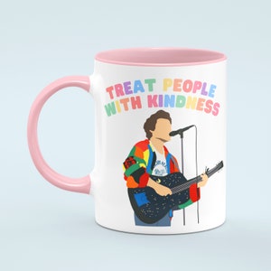 May include: White ceramic mug with a pink rim and handle. The mug features a colourful illustration of a person holding a guitar and singing into a microphone. The text "Treat People With Kindness" is printed in rainbow colours on the mug.