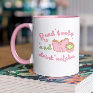 May include: A white ceramic mug with a pink handle and interior. The front features a graphic of an open pink book and a green matcha latte with heart-shaped foam, accompanied by the pink text "Read books and drink matcha" with small stars.