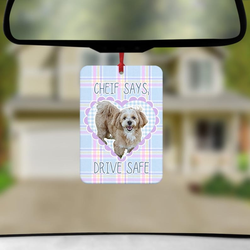 May include: A rectangular car air freshener hanging from a rearview mirror. The design features a light blue, pink, and white plaid background with a central purple heart frame containing a tan dog. Text reads: CHEIF SAYS, DRIVE SAFE.