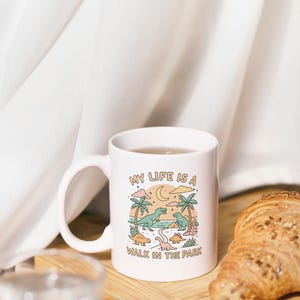 May include: White ceramic mug with a whimsical illustration of dinosaurs in a park setting, featuring the text "MY LIFE IS A WALK IN THE PARK." The mug is next to a croissant on a wooden board.
