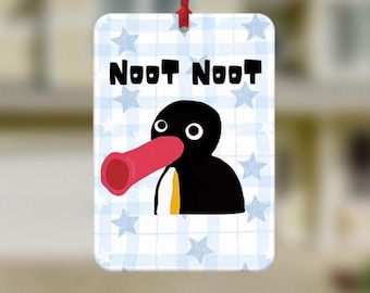 May include: A rectangular ornament featuring a cartoon penguin with a red beak and the words "Noot Noot" in black. The background is a light blue and white checkered pattern with blue stars. A red ribbon is attached for hanging.