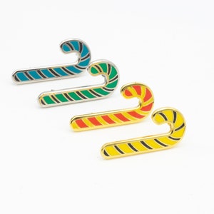 Op de afbeelding: Vier snoepstok-spelden in verschillende kleuren: blauw, groen, rood en geel. Elke speld is gemaakt van metaal en heeft een gouden afwerking.