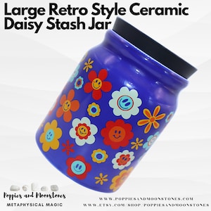 Op de afbeelding: Een grote blauwe keramische pot met een zwart deksel. De pot is versierd met een retro madeliefjespatroon in tinten oranje, geel, rood en wit. De pot is schuin gekanteld en het deksel is lichtjes open.