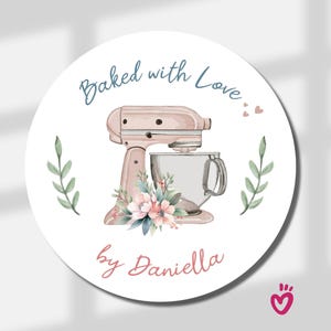 May include: A circular sticker featuring a light pink stand mixer with a glass bowl, decorated with a floral arrangement at the base. The text reads Baked with Love at the top and by Daniella at the bottom, flanked by two green leaf sprigs.