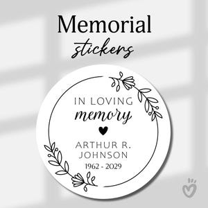 May include: A round white memorial sticker featuring a black line art floral wreath. The text reads "IN LOVING memory" with a small heart icon, followed by the name "ARTHUR R. JOHNSON" and the years "1962 - 2029" printed in a clean sans-serif font.