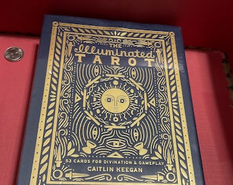 May include: A navy blue tarot card deck titled "The Illuminated Tarot" with gold foil accents. The box features intricate illustrations, including a sun with a face. The text reads "53 Cards for Divination & Gameplay" and the author's name, Caitlin Keegan.