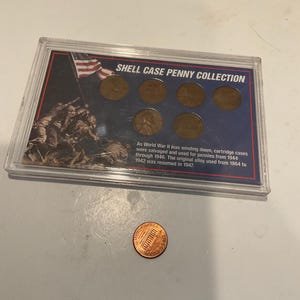May include: A Shell Case Penny Collection display case with six pennies and a single penny. The display case has a graphic of soldiers and an American flag. Text on the case reads "SHELL CASE PENNY COLLECTION".