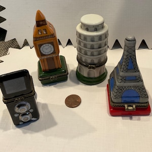 May include: A collection of miniature trinket boxes shaped like famous landmarks: Big Ben, the Leaning Tower of Pisa, the Eiffel Tower, and a vintage camera. The boxes are painted in various colors and sit on a white surface.