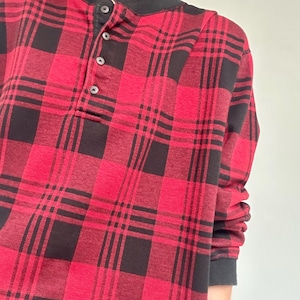 Puede incluir: Una camisa Henley a cuadros rojos y negros con cuello y puños negros. La camisa tiene una tapeta abotonada con tres botones. La camisa de manga larga está hecha de una tela suave y texturizada.