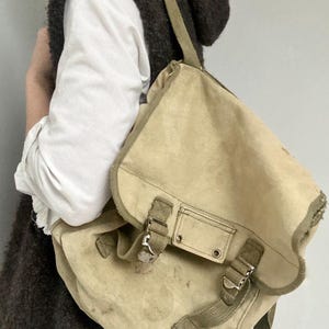 May include: A tan canvas messenger bag with olive green trim and straps. The bag features a flap closure with two metal buckles and a small pocket. The bag is worn over the shoulder.