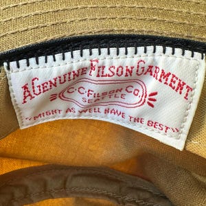 May include: A tan cap featuring an interior white fabric label with red embroidered text that reads A GENUINE FILSON GARMENT, C.C. FILSON CO. SEATTLE, and MIGHT AS WELL HAVE THE BEST, stitched onto a black band inside the crown of the hat.