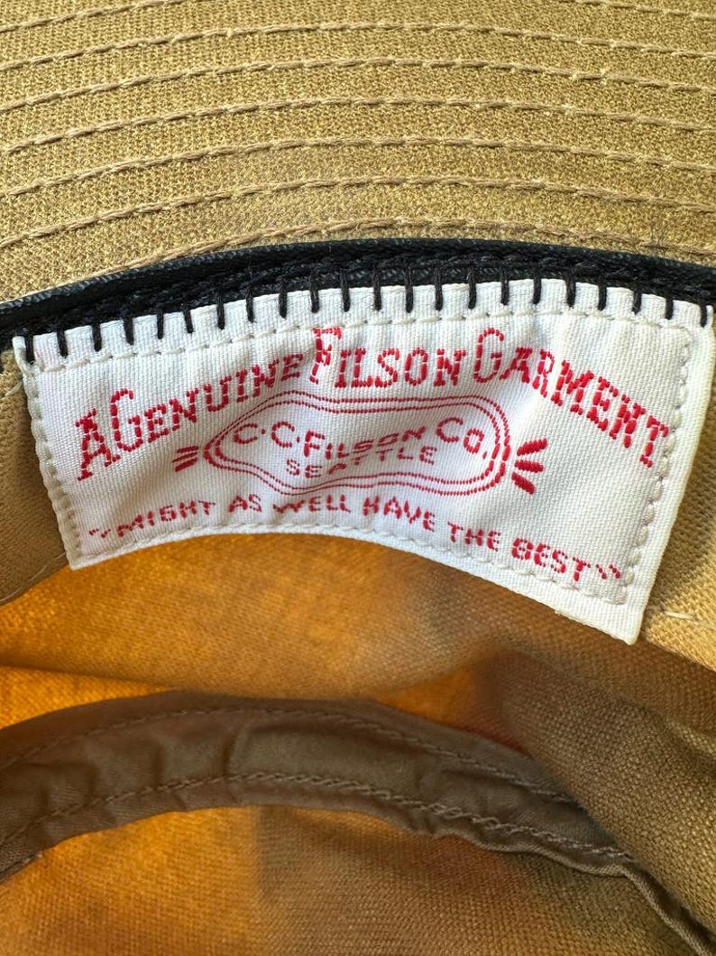 May include: A tan cap featuring an interior white fabric label with red embroidered text that reads A GENUINE FILSON GARMENT, C.C. FILSON CO. SEATTLE, and MIGHT AS WELL HAVE THE BEST, stitched onto a black band inside the crown of the hat.