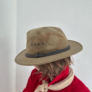May include: An olive green fedora hat with a dark brown leather band and four small metal ventilation eyelets on the side. The hat features a structured crown and a wide brim, providing a classic look with a durable, matte fabric finish.