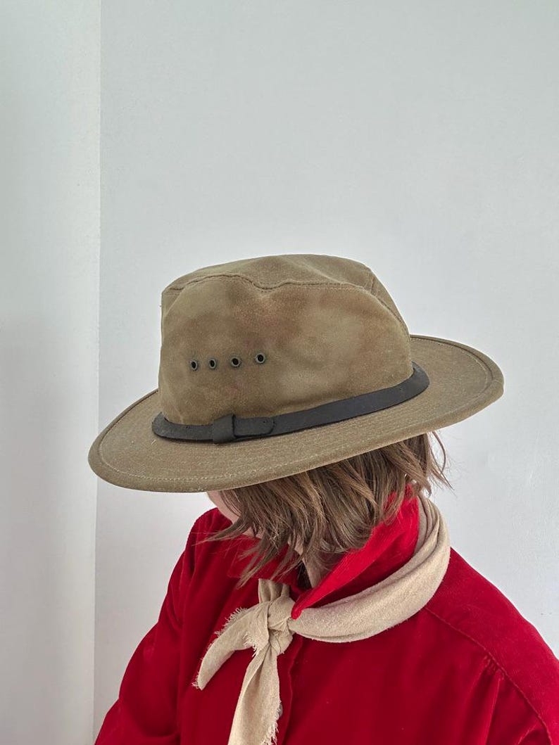 May include: An olive green fedora hat with a dark brown leather band and four small metal ventilation eyelets on the side. The hat features a structured crown and a wide brim, providing a classic look with a durable, matte fabric finish.