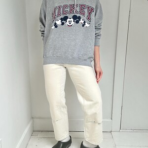 May include: A gray Mickey Mouse hoodie with a navy blue hood and the word "MICKEY" in red lettering. The hoodie is paired with cream-colored jeans and black clogs. The outfit is casual and comfortable.