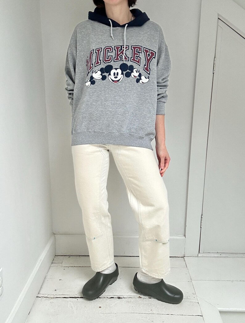 May include: A gray Mickey Mouse hoodie with a navy blue hood and the word "MICKEY" in red lettering. The hoodie is paired with cream-colored jeans and black clogs. The outfit is casual and comfortable.