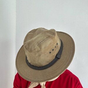 May include: A tan fedora-style hat with a wide brim and a dark brown band around the crown. The hat features four small metal ventilation eyelets on the side. The hat is worn over a light-colored fabric piece, positioned above a bright red shirt.