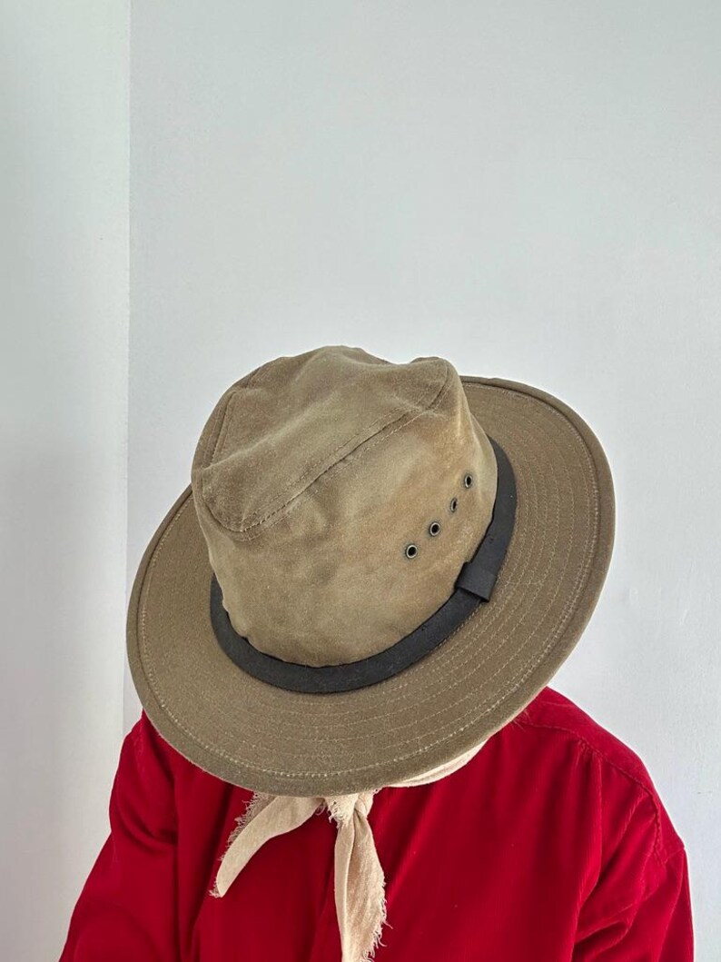May include: A tan fedora-style hat with a wide brim and a dark brown band around the crown. The hat features four small metal ventilation eyelets on the side. The hat is worn over a light-colored fabric piece, positioned above a bright red shirt.