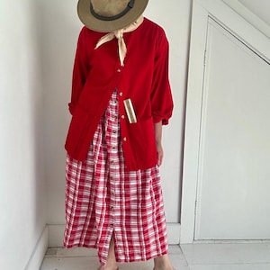 May include: A bright red button-down long-sleeve shirt with two front pockets, worn over a red and white plaid button-down midi dress. The outfit includes a tan fedora hat, a light beige neck scarf, and tan slide sandals with wide straps.