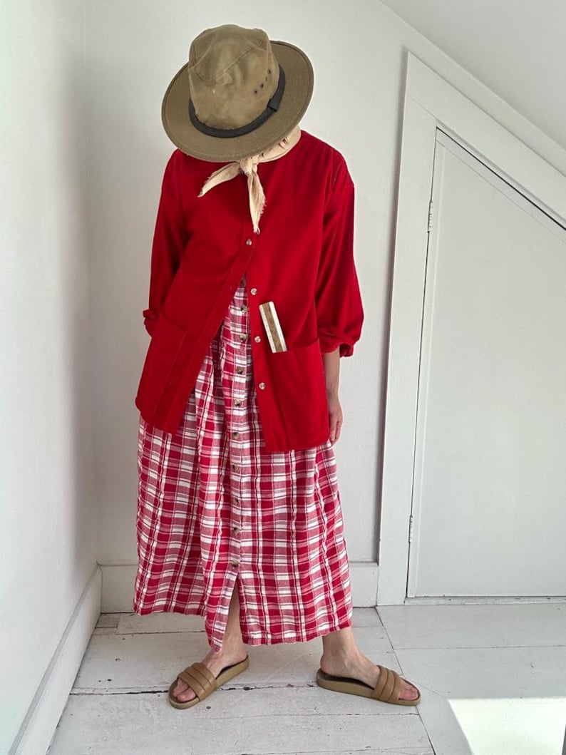 May include: A bright red button-down long-sleeve shirt with two front pockets, worn over a red and white plaid button-down midi dress. The outfit includes a tan fedora hat, a light beige neck scarf, and tan slide sandals with wide straps.