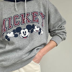 May include: A gray hooded sweatshirt featuring the word "MICKEY" in maroon lettering with a white outline. Below the text are three Mickey Mouse character illustrations. The hood is navy blue with a white drawstring.