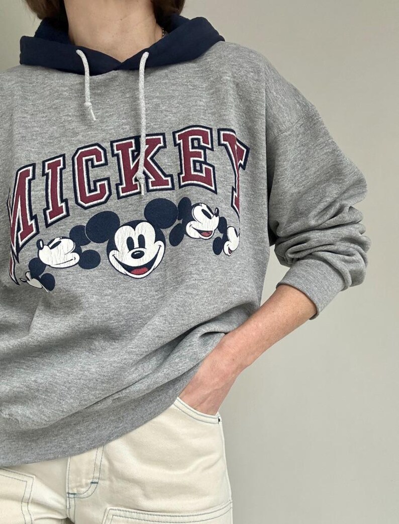 May include: A gray hooded sweatshirt featuring the word "MICKEY" in maroon lettering with a white outline. Below the text are three Mickey Mouse character illustrations. The hood is navy blue with a white drawstring.